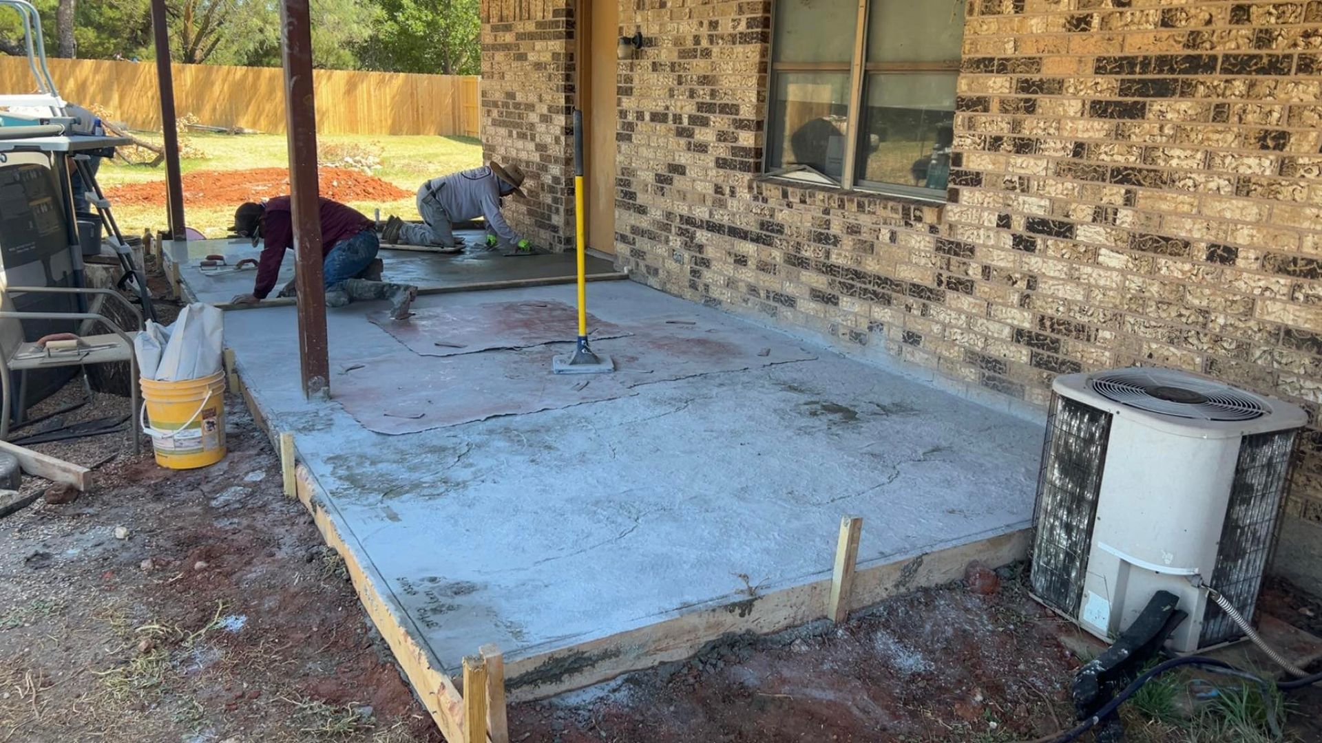 Concrete patio being poured next to brick building; workers spread concrete with tools.