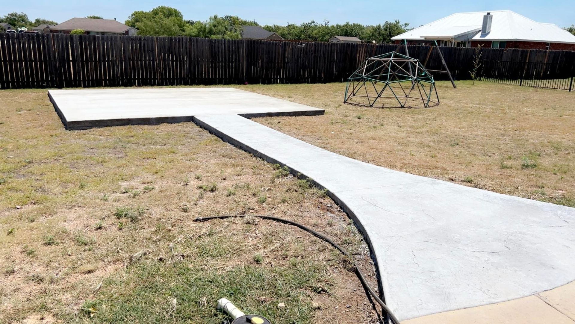 Concrete path and pad in a grassy backyard with a play structure and fence.