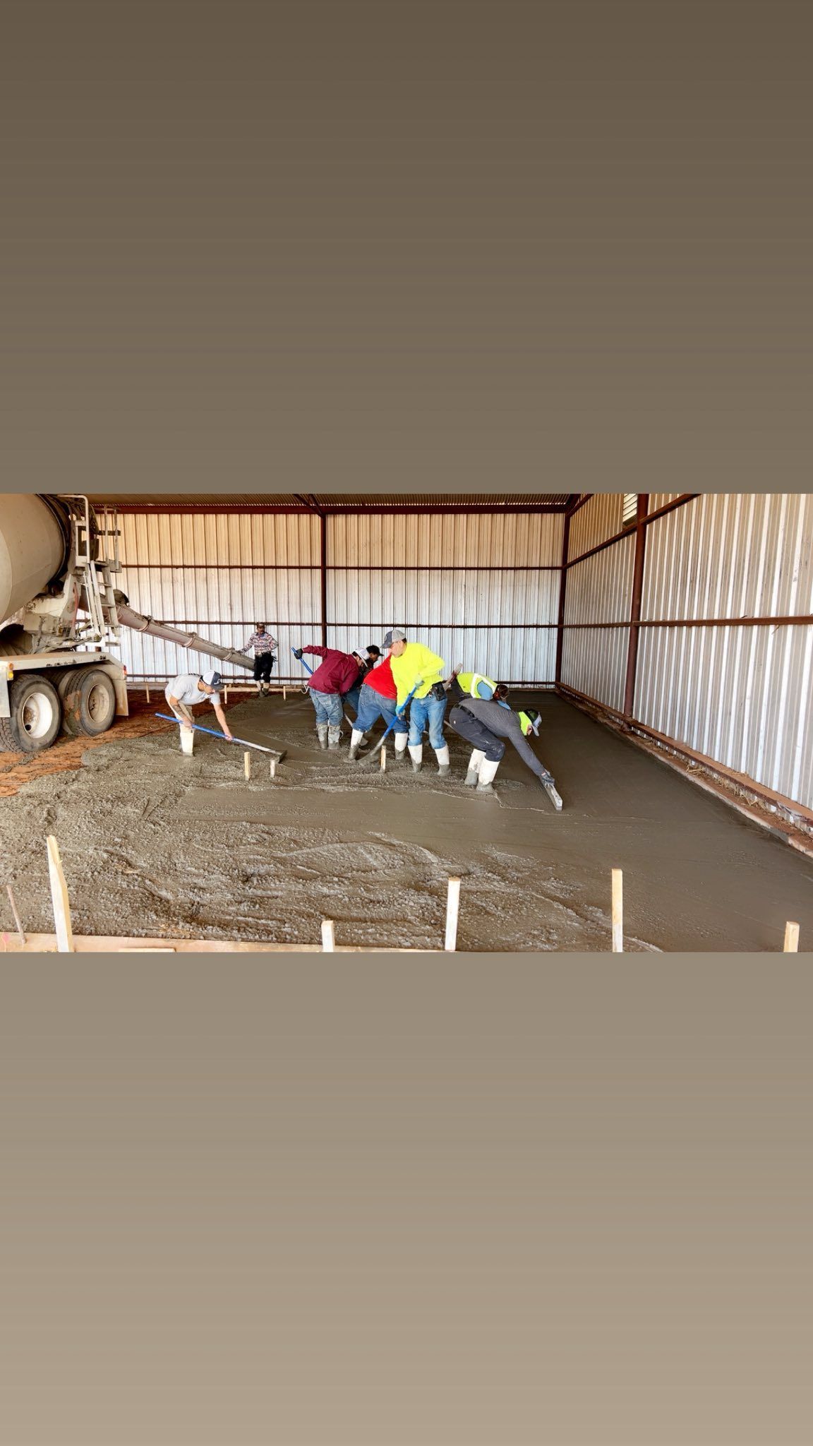 Workers pouring concrete floor inside a metal building; concrete truck visible, leveling the surface.