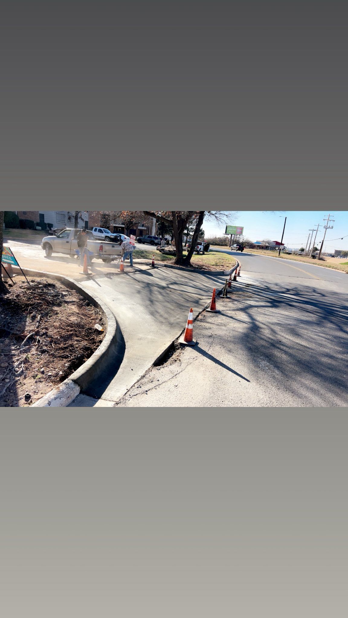 Road construction. Concrete sidewalk, asphalt street, orange cone, workers in background.
