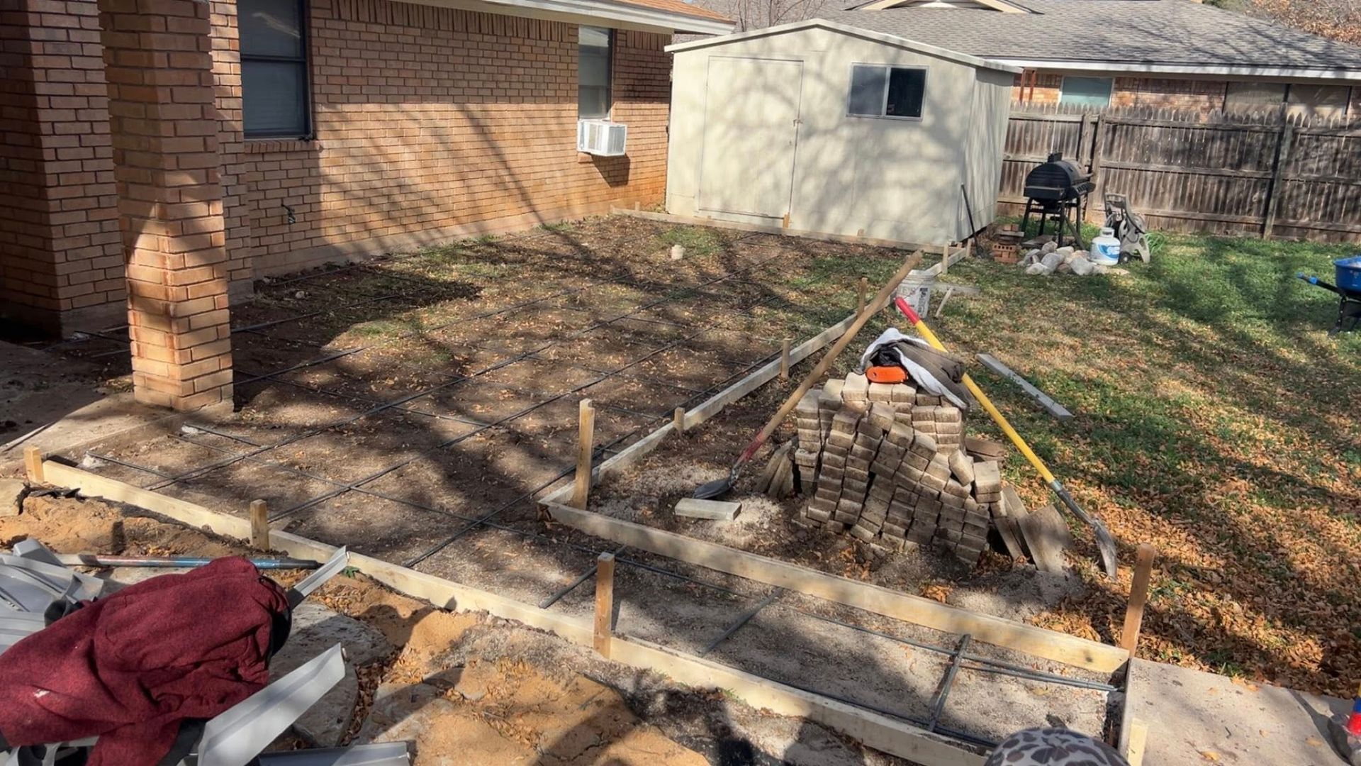 Construction site: concrete forms, dirt, brick building, shed, tree stump, and tools.