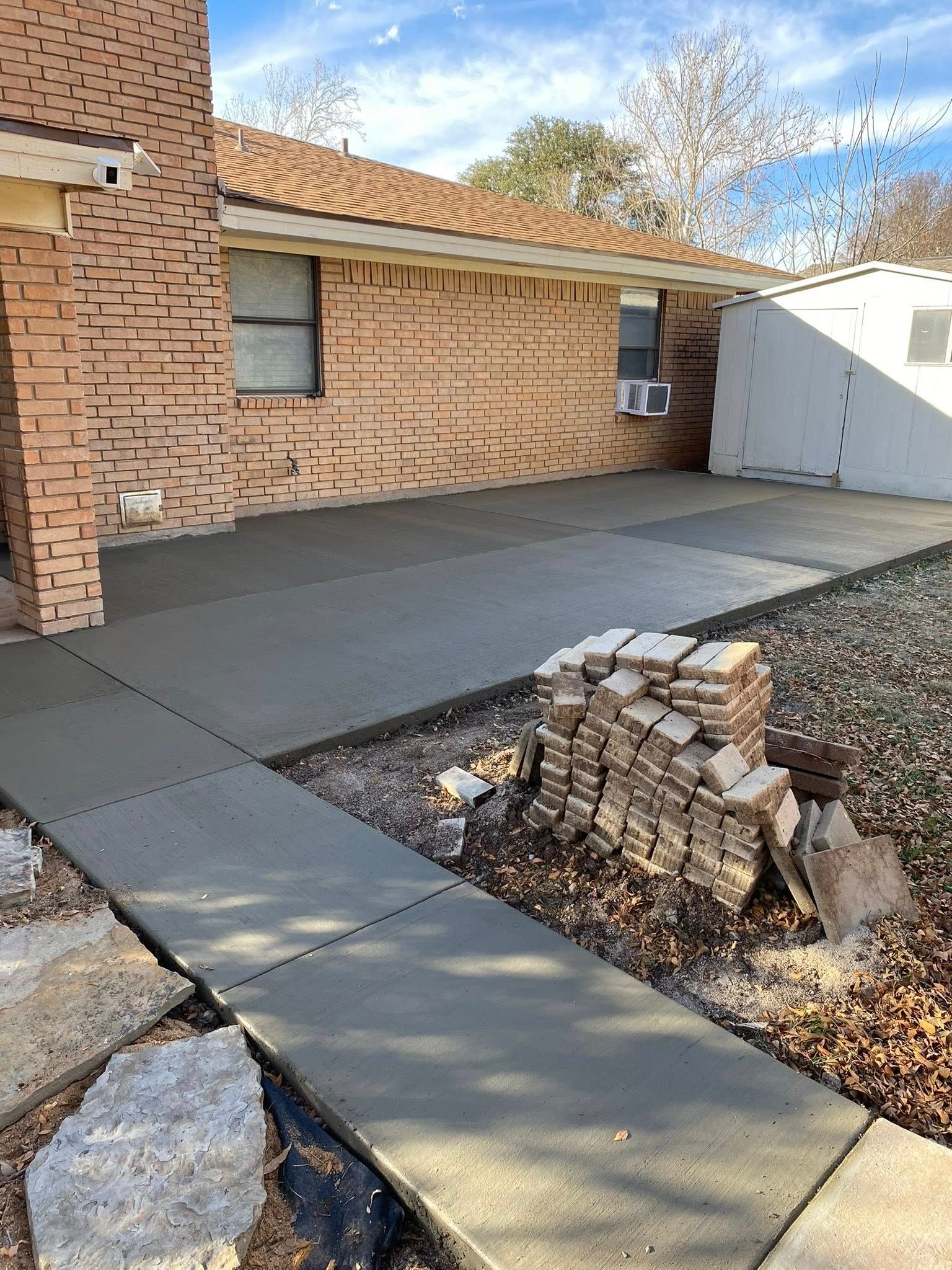 Newly poured concrete patio and sidewalk next to a brick building, with a few rocks and dry vegetation.