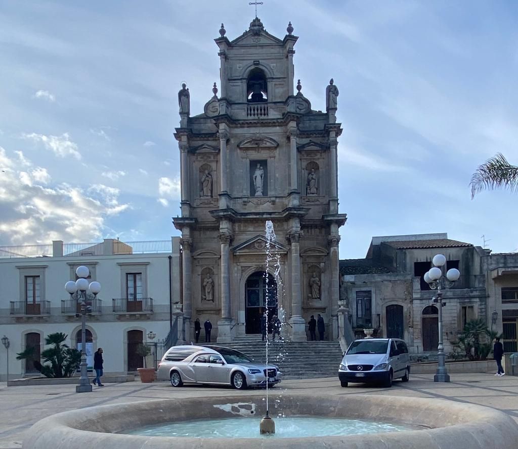 Facciata barocca della chiesa con fontana, automobili e piazza. Cielo luminoso.