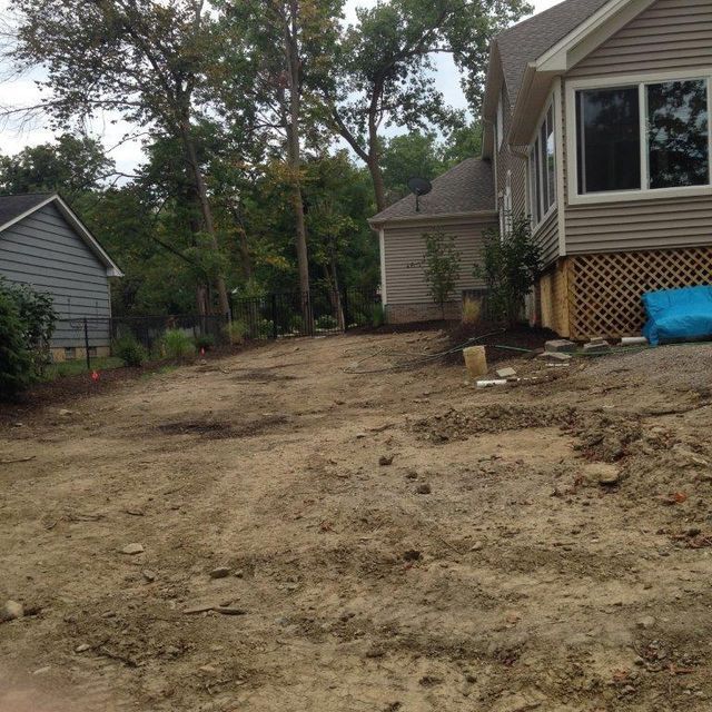 Dirt driveway beside a house under construction, with trees and neighboring homes in the background