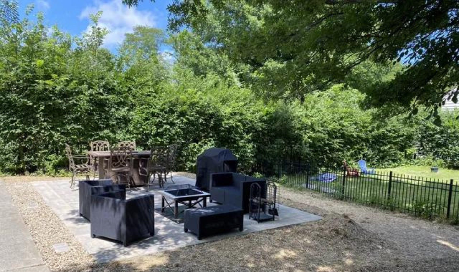 Outdoor patio with black grill and seating on gravel, surrounded by trees and a metal fence