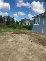 Dirt yard beside a gray house, with trees and neighboring homes under a blue, cloudy sky