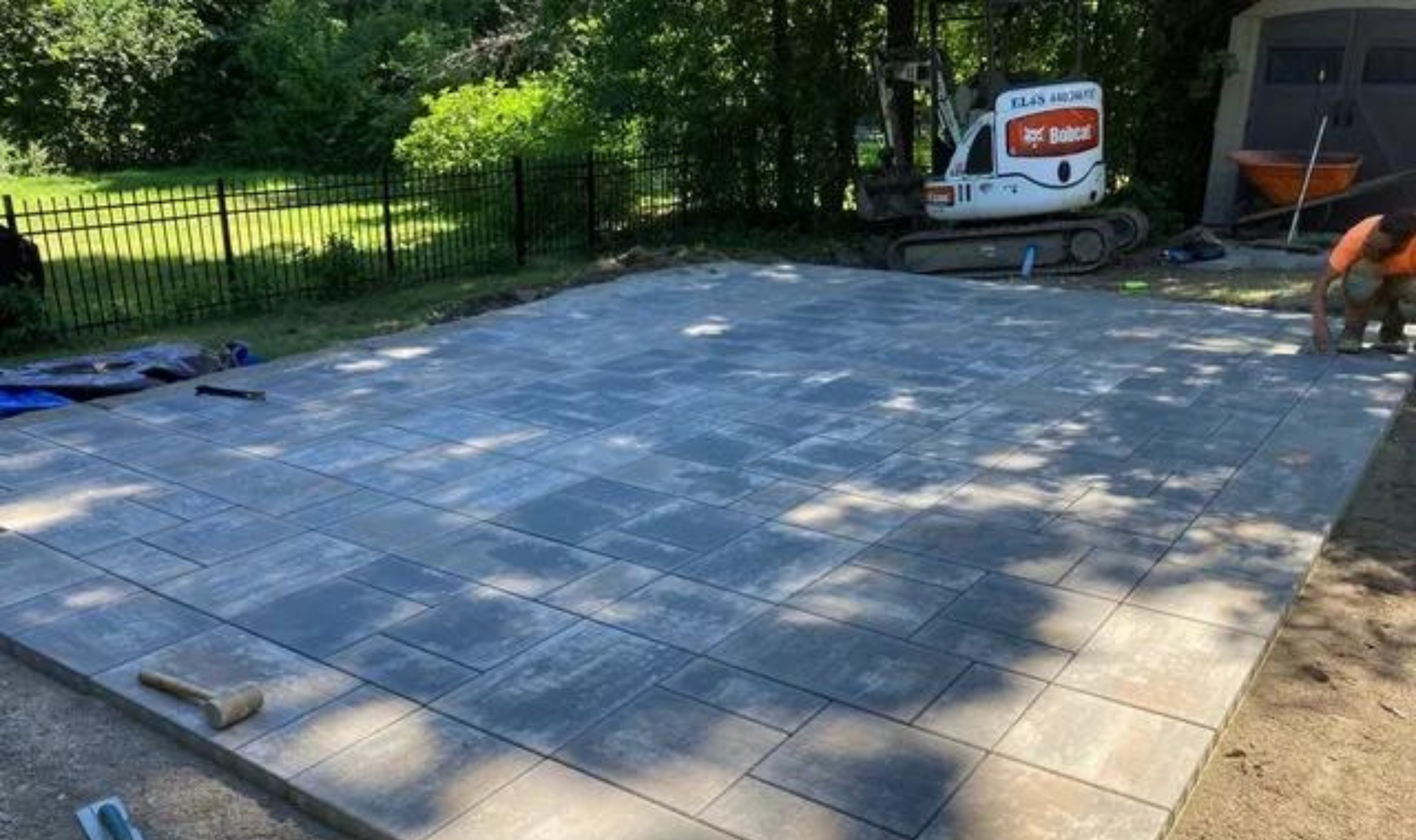 Stone patio under construction with gray pavers, bordered by grass and trees, with a wheelbarrow nearby