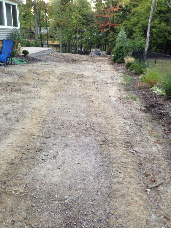 Dirt driveway under construction beside trees and shrubs, with gravel and equipment along the edges