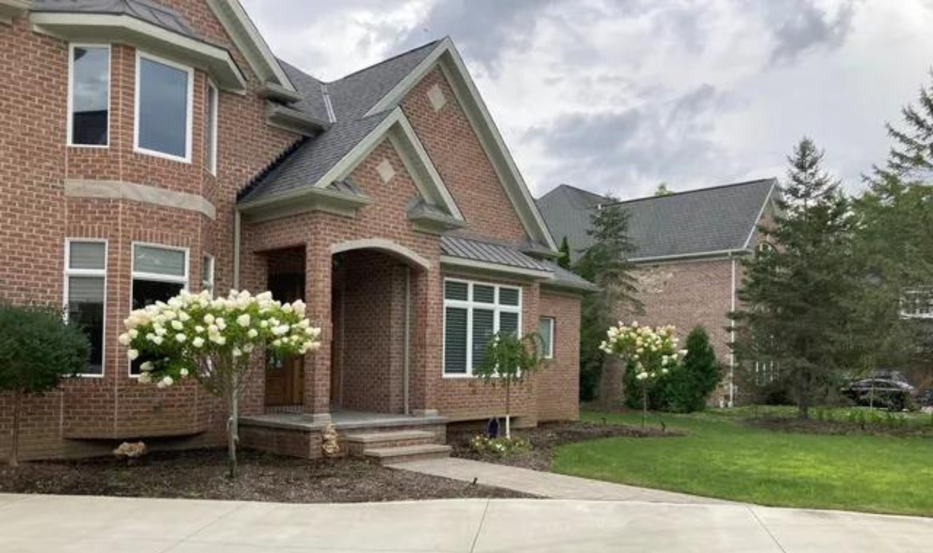 Brick suburban house with covered front porch, white flowering shrubs, and a manicured lawn.
