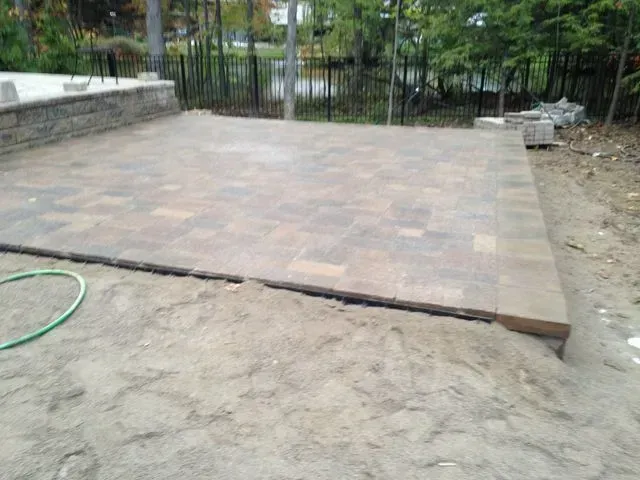 Raised tiled patio beside a dirt yard, with a metal fence and trees in the background.