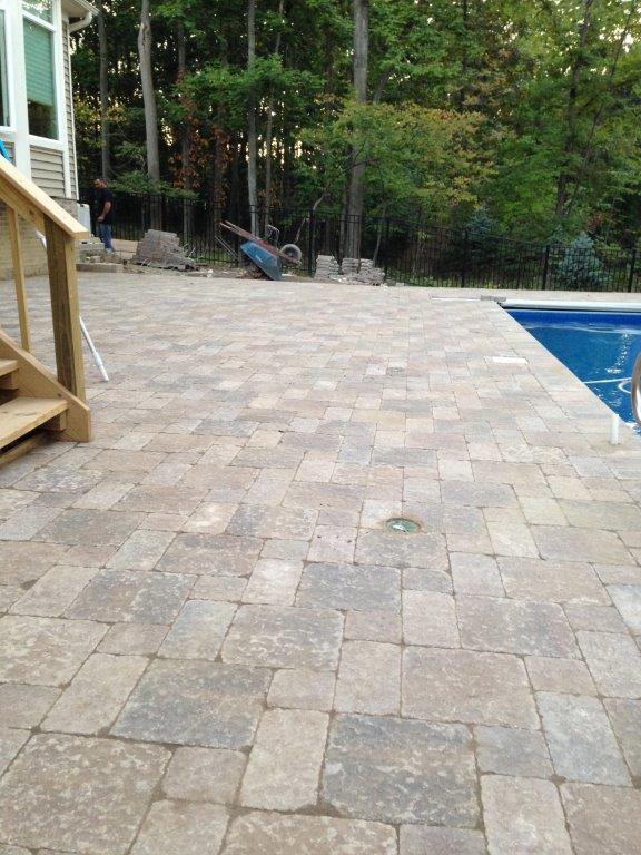 Stone patio beside a blue swimming pool, bordered by trees and a wooden deck railing.