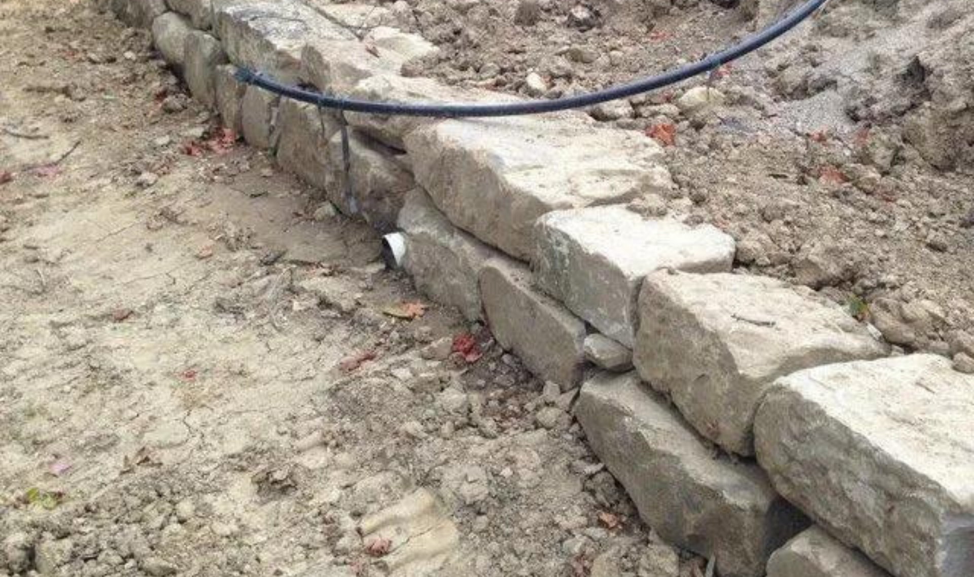 Stone retaining wall under construction beside dirt, with a black hose running along the top.