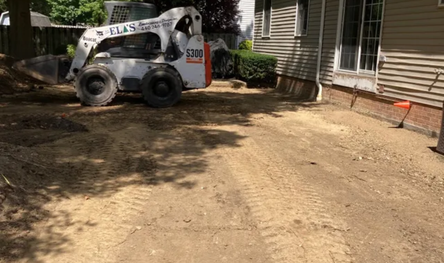 White skid-steer parked on dirt beside a house, with a cleared yard area and shade trees nearby.
