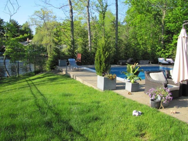 Sunny backyard with green lawn, swimming pool, patio chairs, and potted flowers