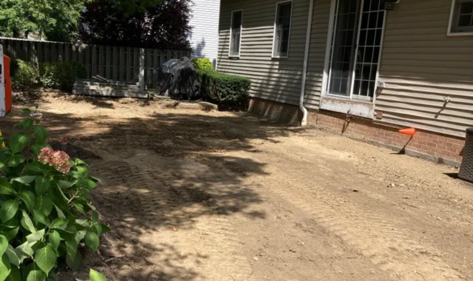 Sunny dirt yard beside a house with sliding glass doors, shaded by trees and bordered by a fence and shrubs