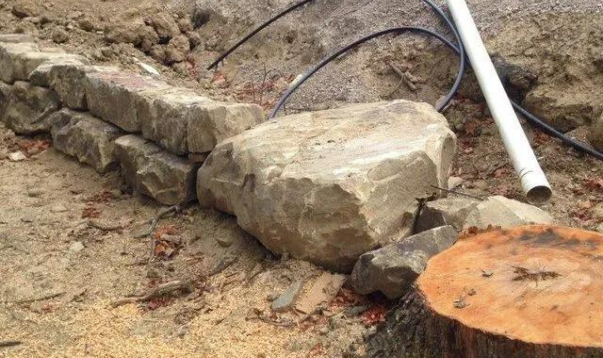 Stone retaining wall under construction beside a large boulder and white pipe in dirt