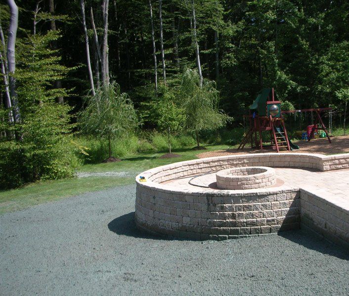 Circular stone fire pit in a wooded park with a playground in the background