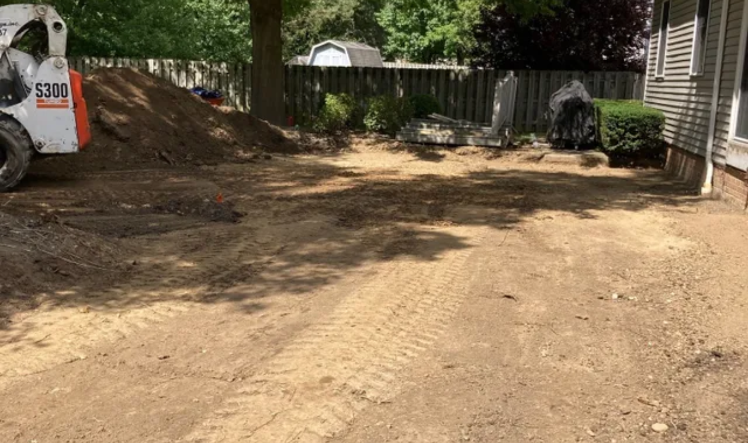 Dirt driveway beside a white vehicle and house, with trees and a fenced yard in the background