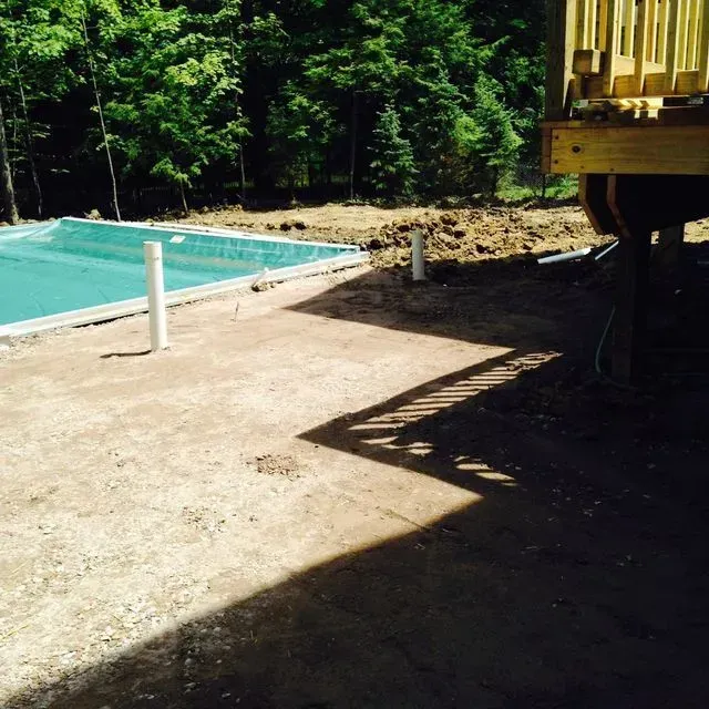 Concrete patio beside a turquoise pool, with forest trees and a wooden structure casting shadows.