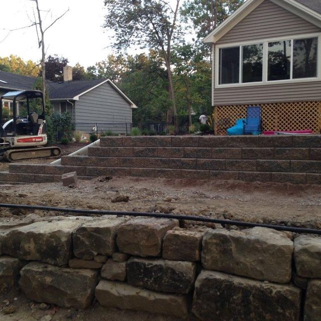 Terraced stone retaining wall and steps in a backyard, with a house and excavator nearby