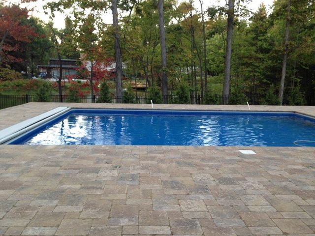 Backyard pool with blue water beside a stone patio and trees in the background