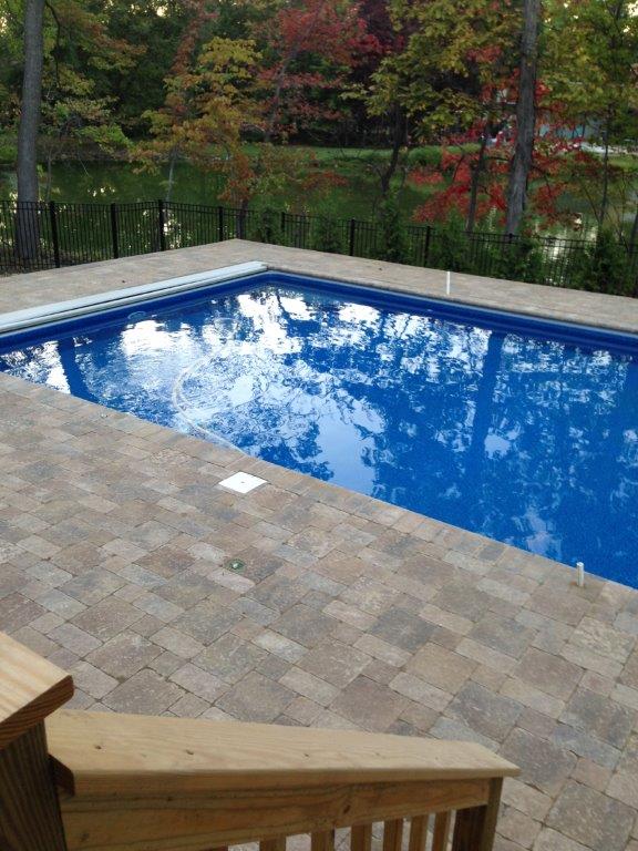 Blue backyard swimming pool beside stone patio and wooden steps, with trees and a lake in the background