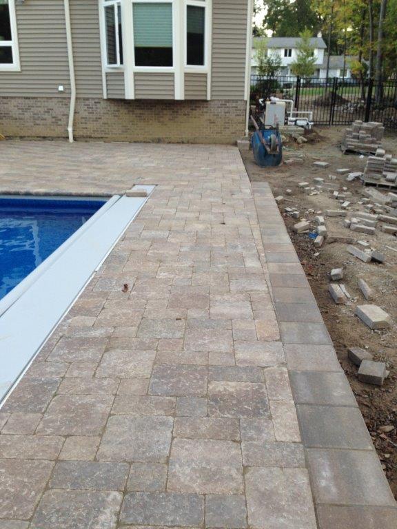 Backyard pool with stone paver patio beside a house under construction, with dirt and loose bricks on the right.