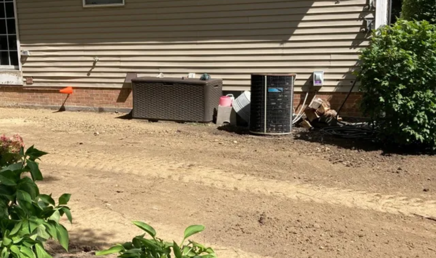 Backyard beside house with dirt yard, HVAC unit, storage bin, and shrubs.