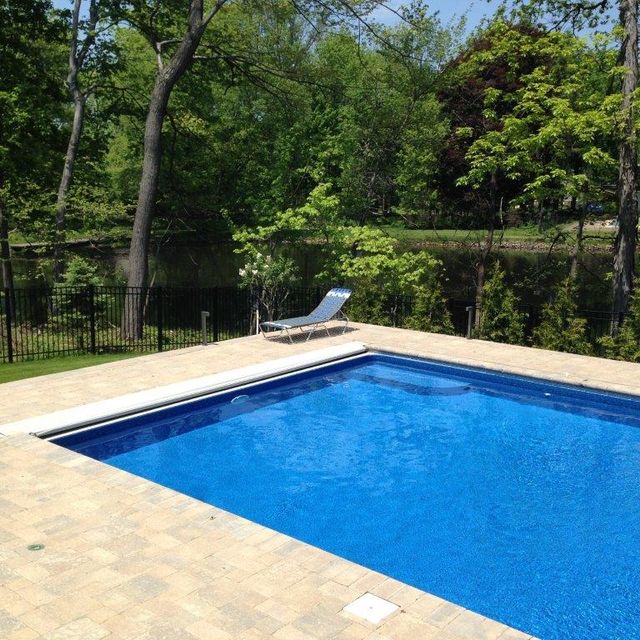 Blue backyard pool beside a tan patio, with a lounge chair and trees reflected in the water