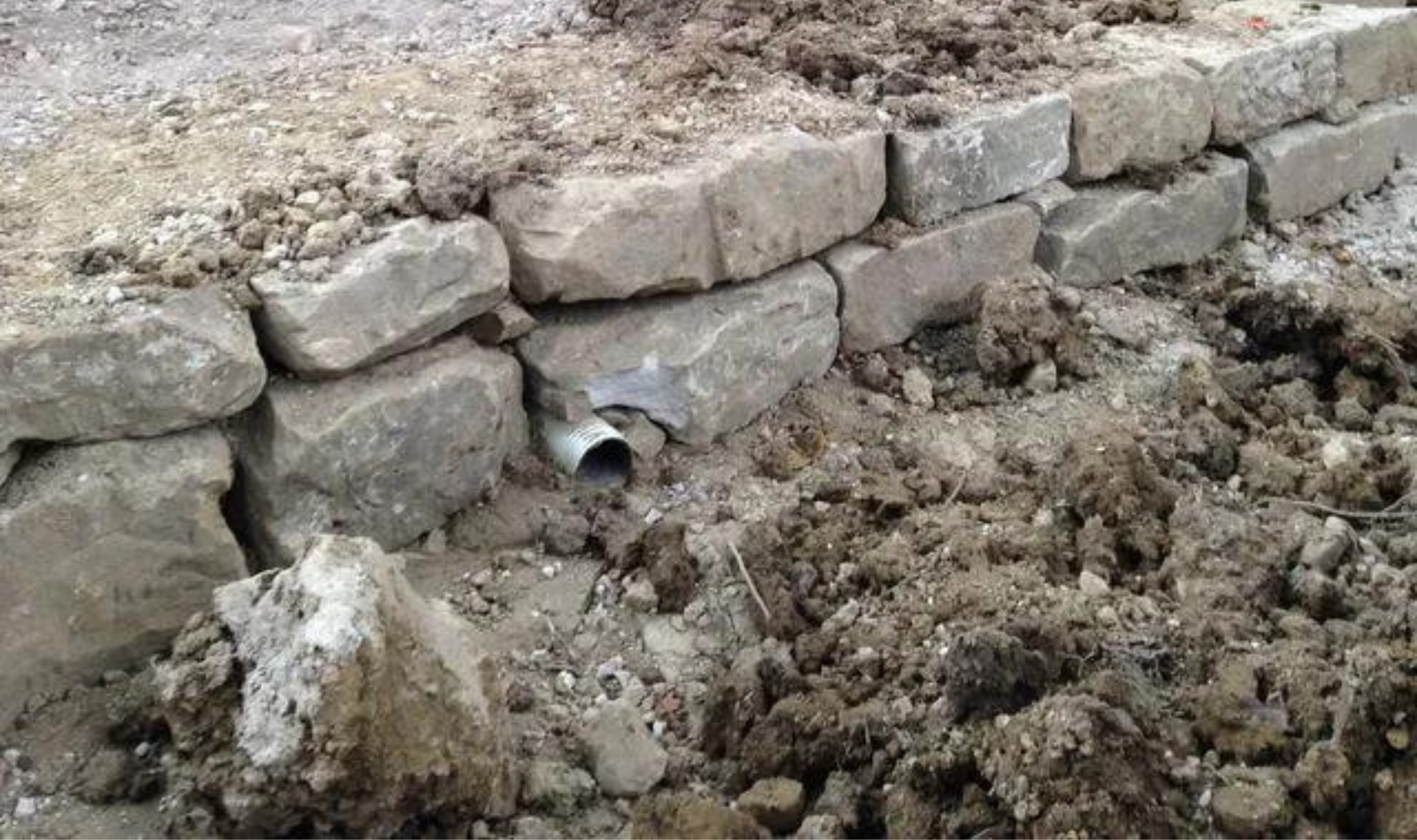 Stone retaining wall with a small pipe outlet, surrounded by dirt and rubble.