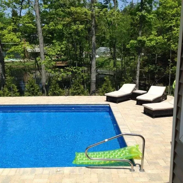 Blue swimming pool with lounge chairs, patio, and trees in the background; pool lift at the edge
