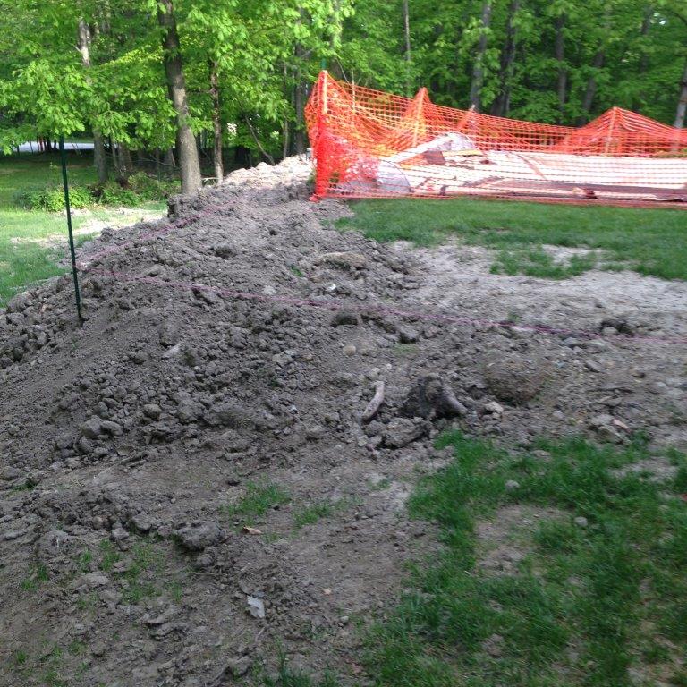 Pile of dirt along a grassy park path beside orange safety fencing and trees.