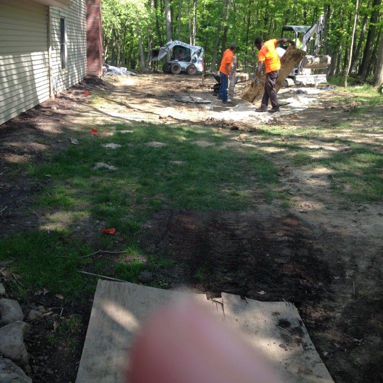 Workers in orange vests using a backhoe in a wooded yard beside a house