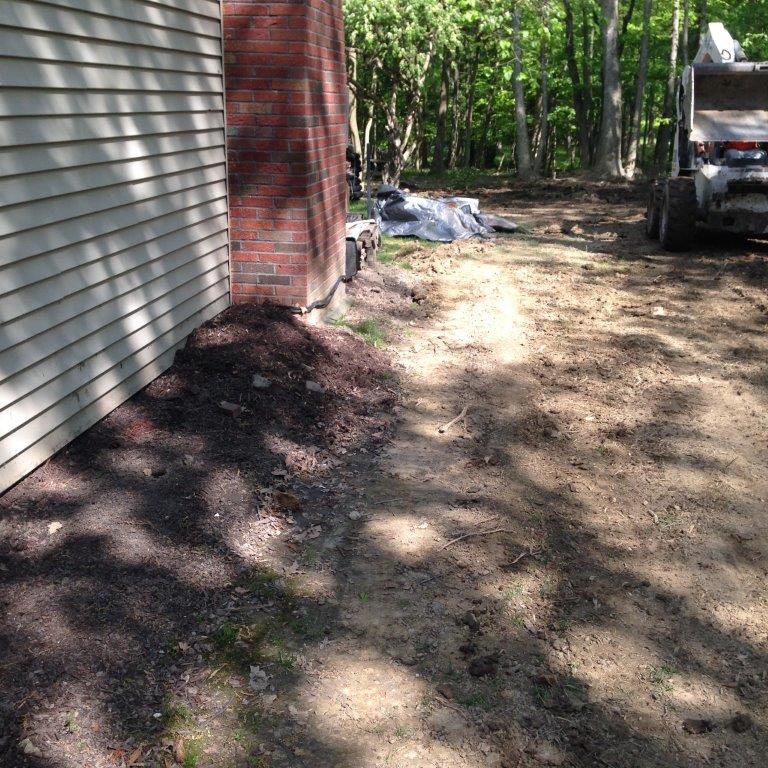 Dirt driveway beside a house, with a truck parked near trees and mulch piles in the yard.