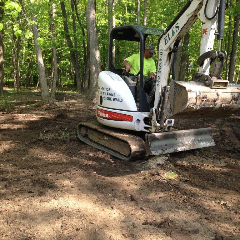 White excavator on muddy ground in a wooded area, with a person operating it