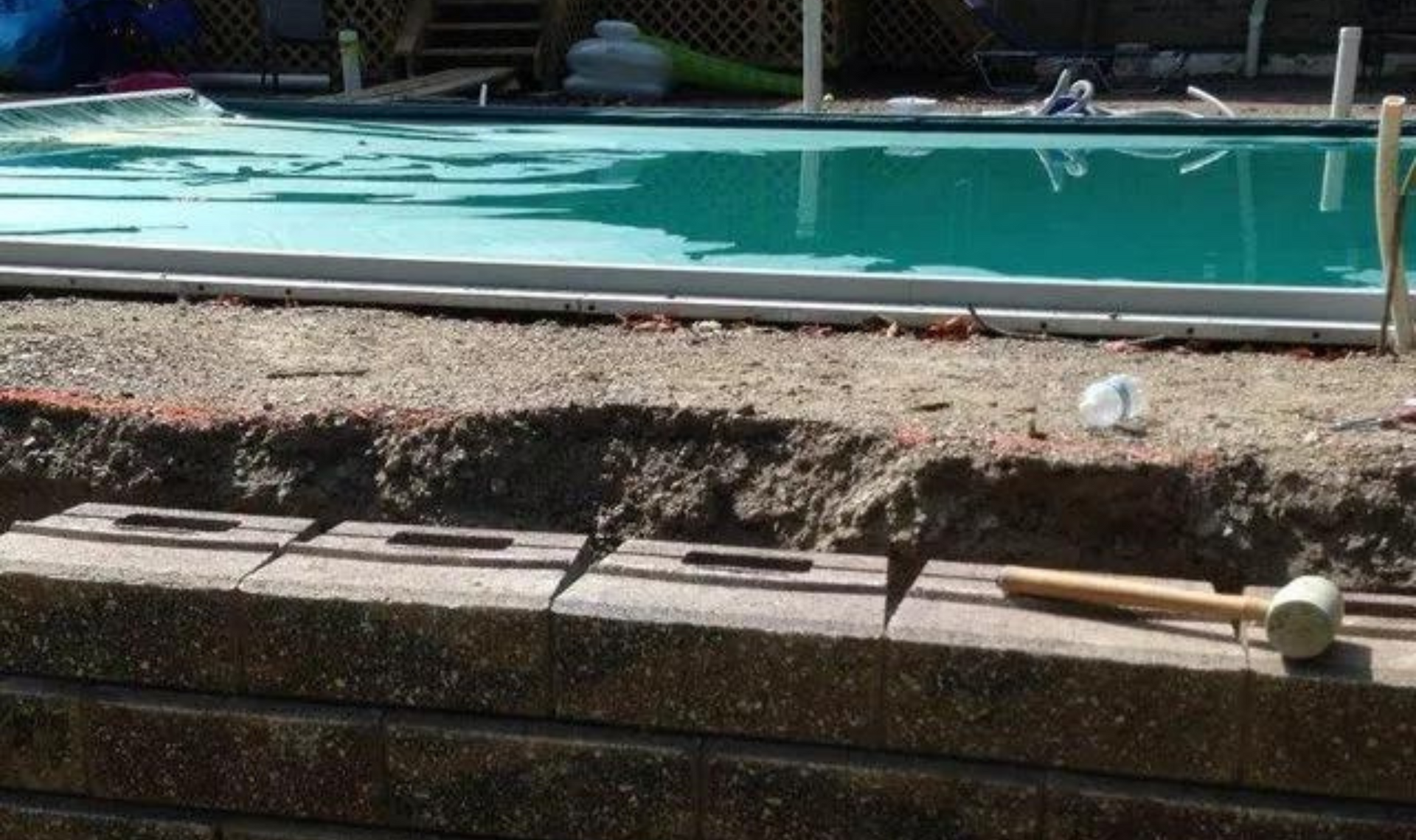 Empty garden planter beside a turquoise swimming pool, with a shovel and a white ball on the ledge.