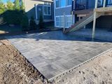 Concrete patio beside a house with steps and a raised deck, bordered by gravel and soil