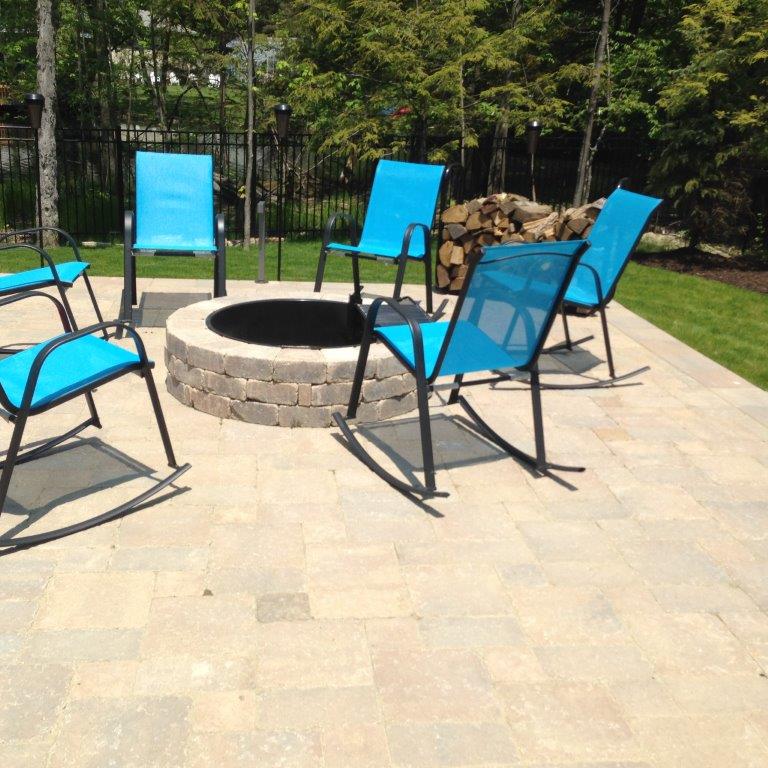 Blue patio chairs around a stone fire pit on a sunlit paved backyard patio.