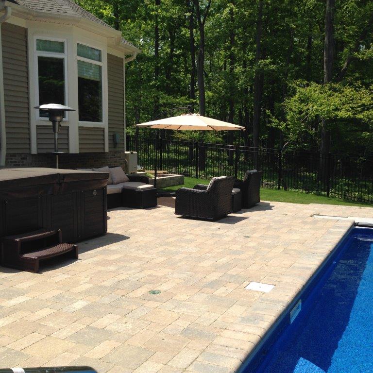 Patio with outdoor seating and umbrella beside a blue swimming pool, backed by trees and a house.