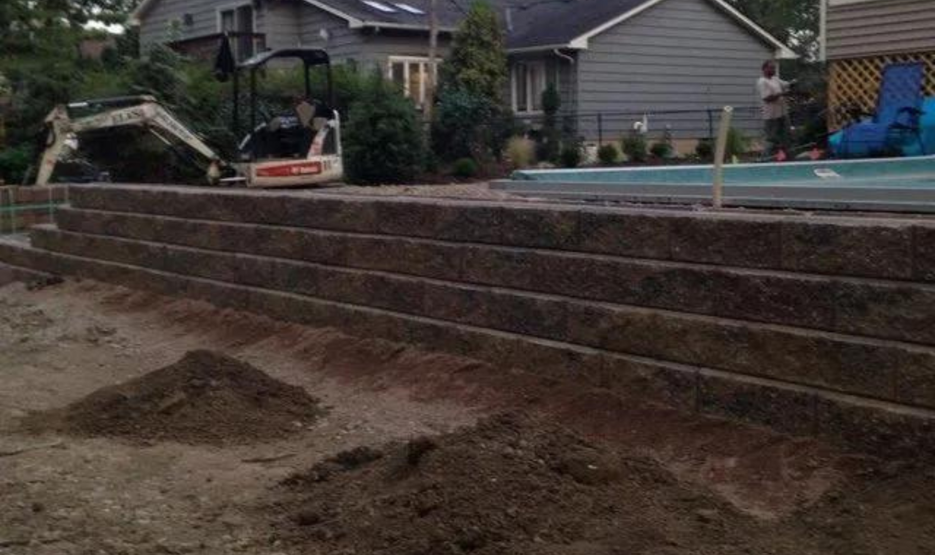 Backyard with tiered stone retaining walls beside a house and a swimming pool, under construction