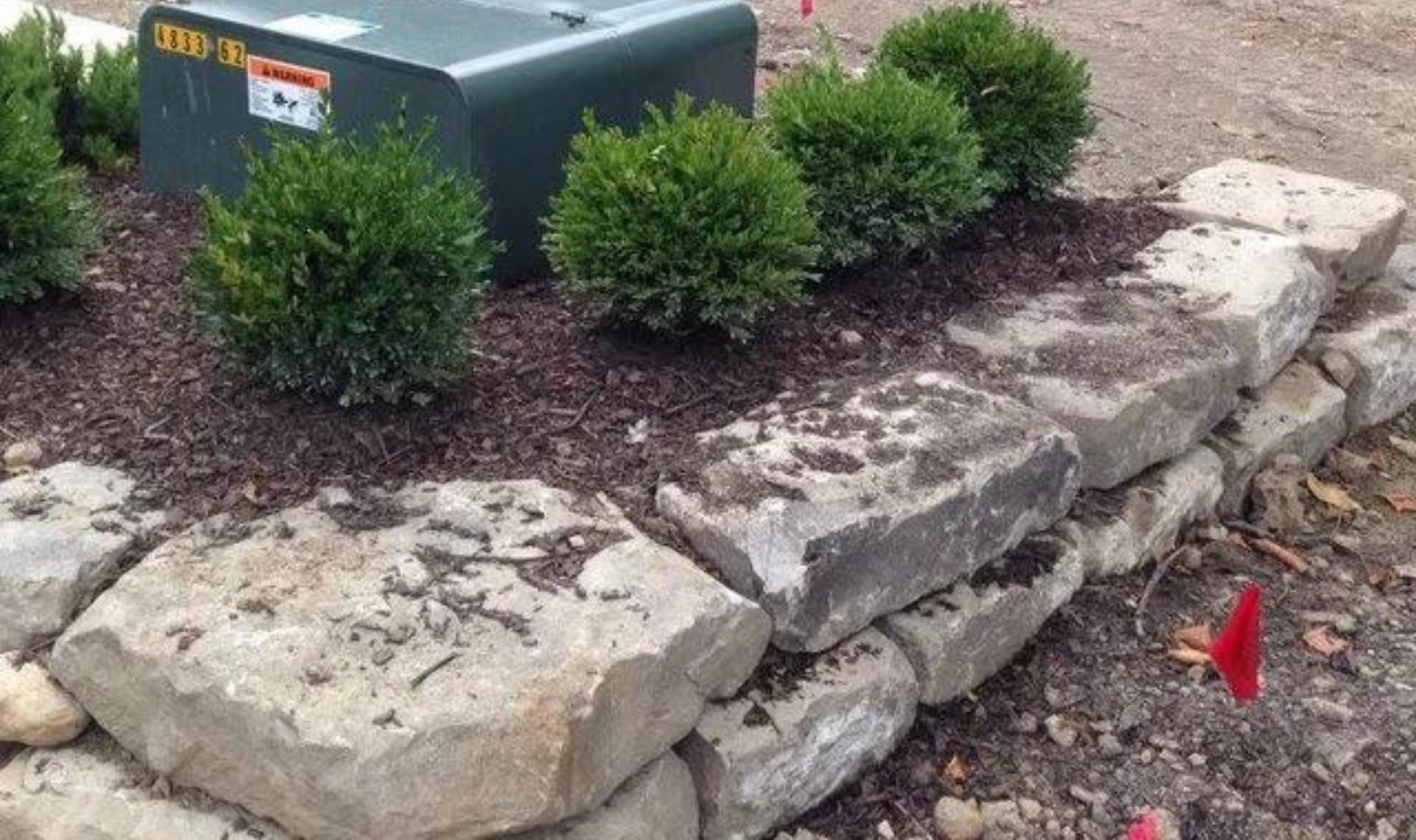Stone retaining wall with small shrubs and a utility box in a landscaped area
