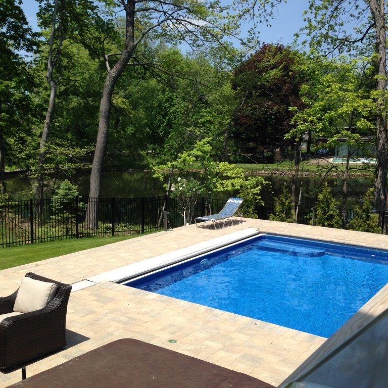 Backyard swimming pool with tan stone deck, lounge chair, and green trees beyond a black fence