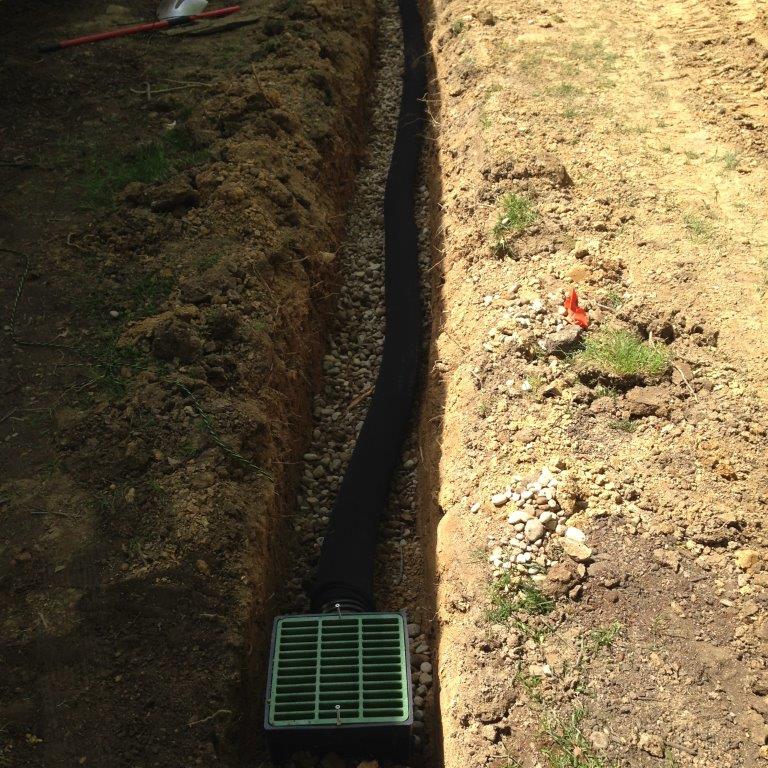 Narrow trench in dirt with a green drain grate at the bottom.