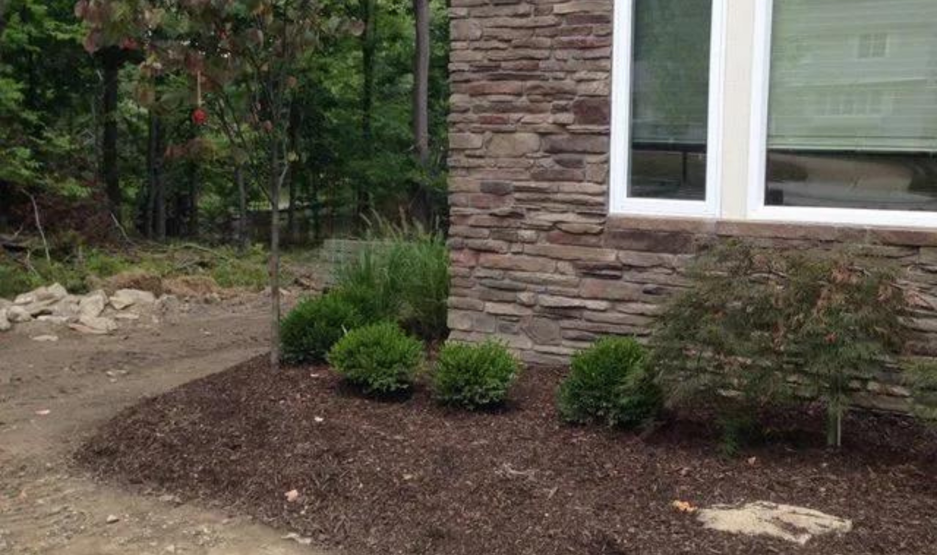 Landscaped yard beside a stone house with shrubs, mulch, and a dirt path through trees.