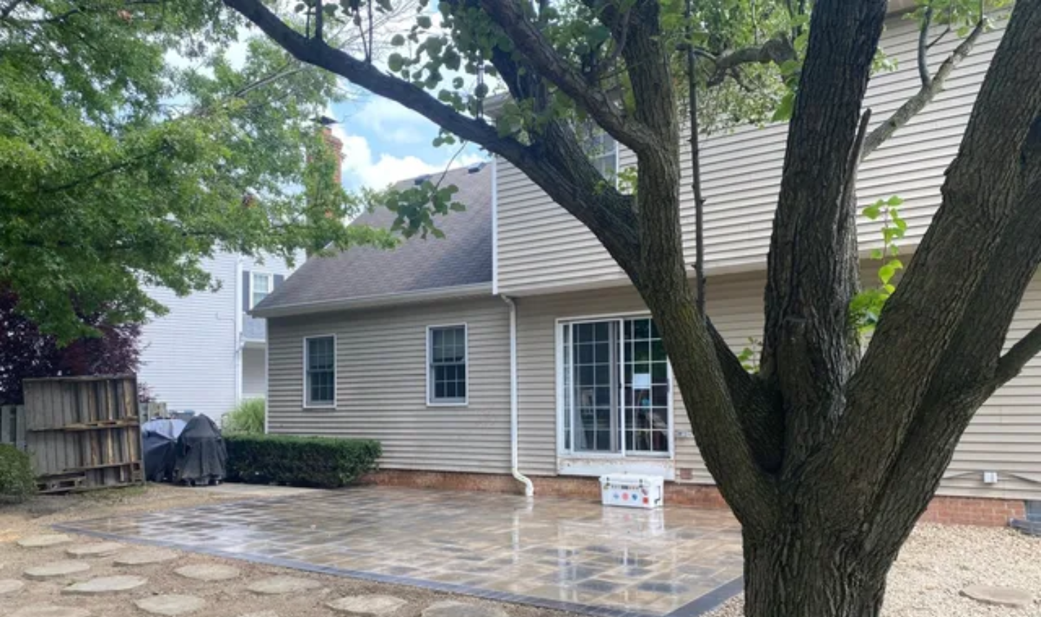 Backyard patio beside a beige house, with a large tree in the foreground and white French doors.