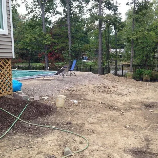 Backyard with a swimming pool, chairs, trees, and a bare dirt area beside a house.