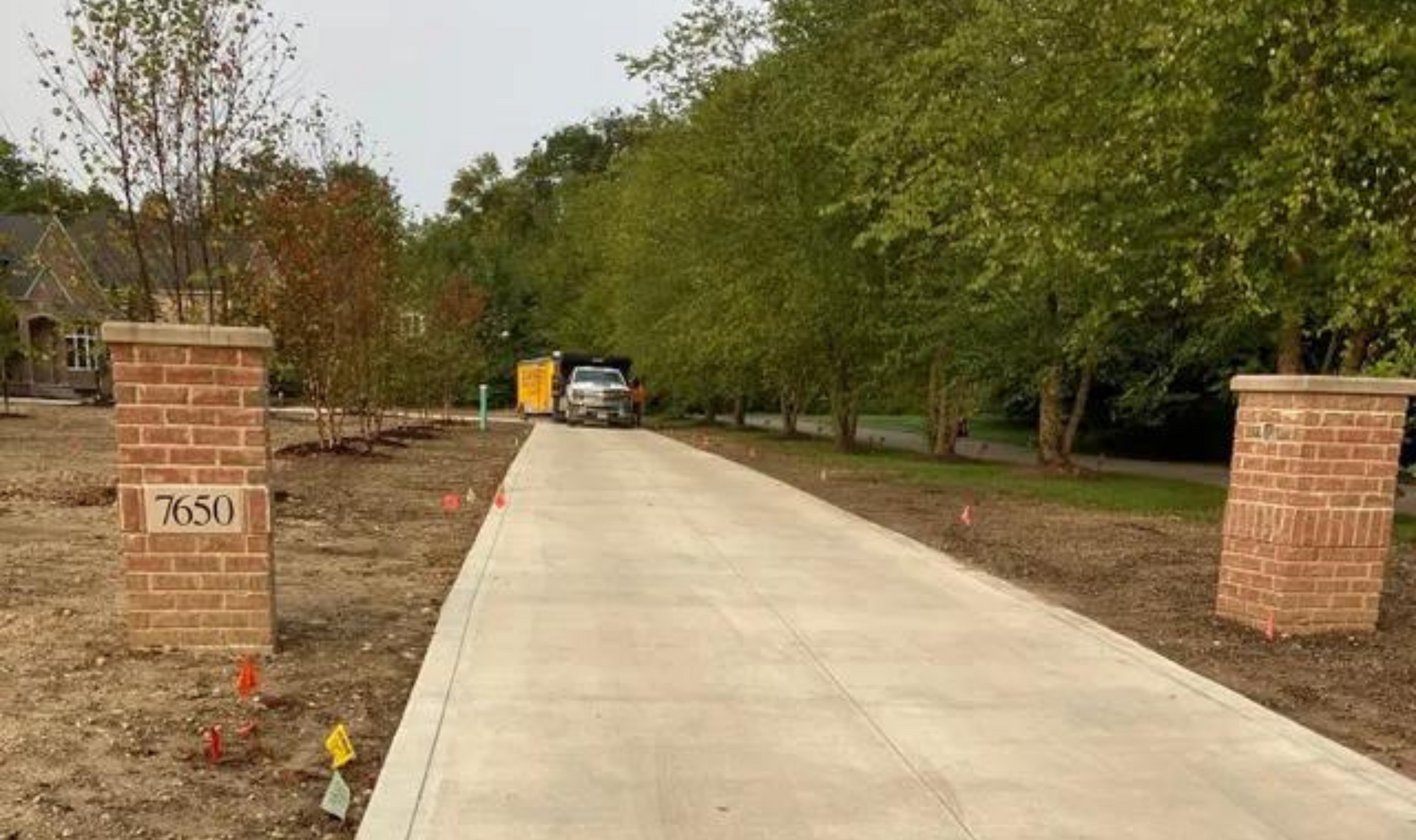 Concrete driveway between brick pillars leading to a yellow vehicle, lined with trees on both sides