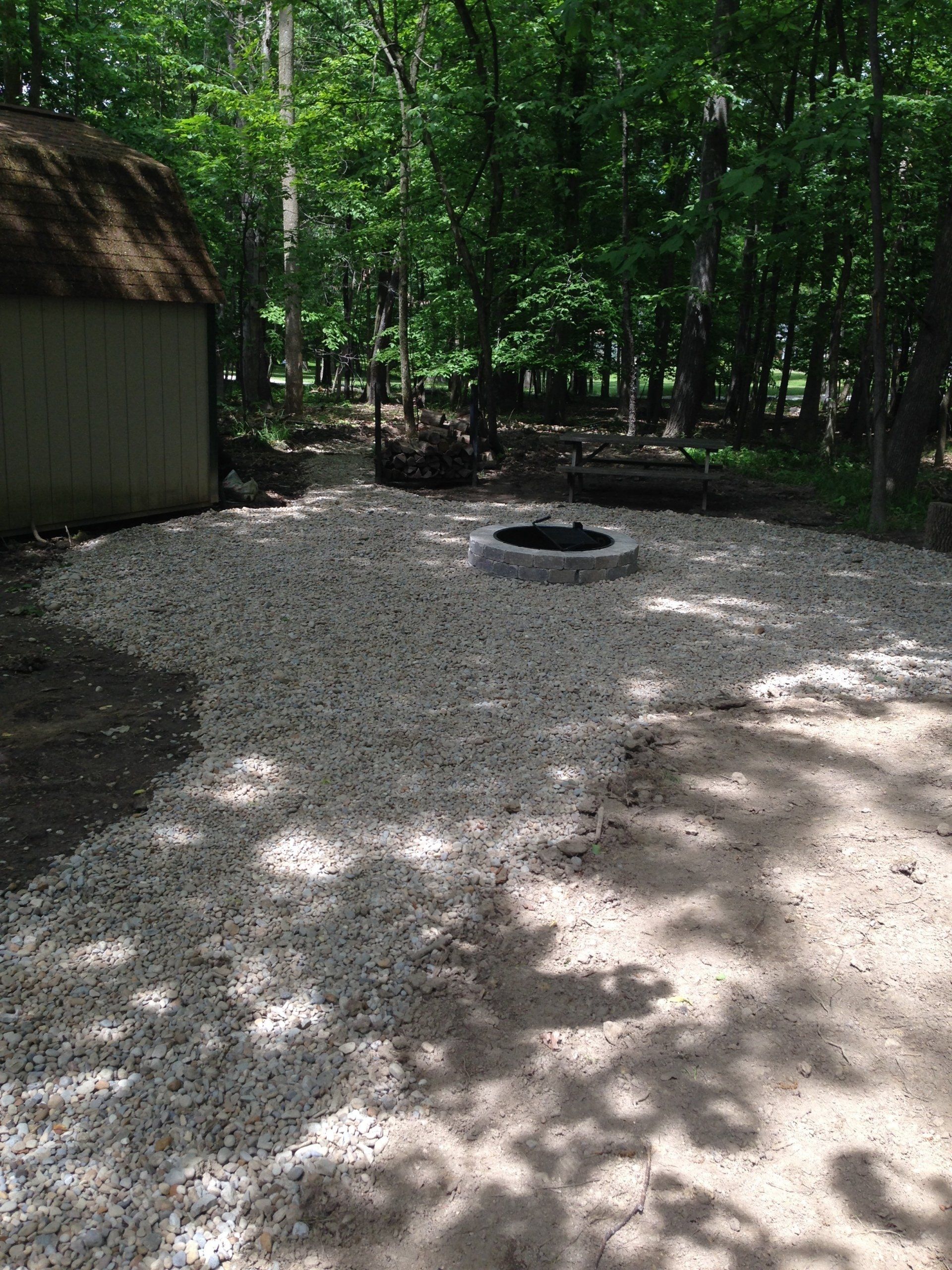 Gravel campsite with a circular fire pit beside a wooded forest and a small building.