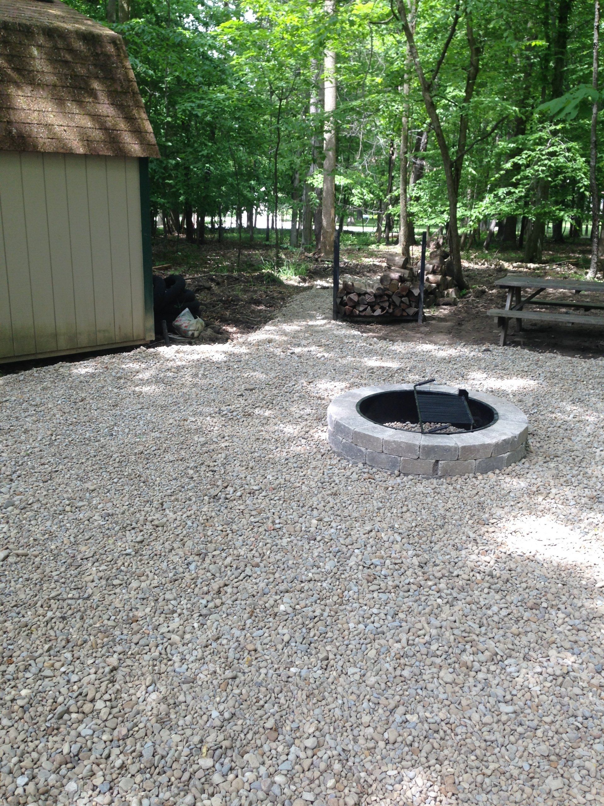 Gravel campsite with circular fire pit beside a small shed in a wooded area
