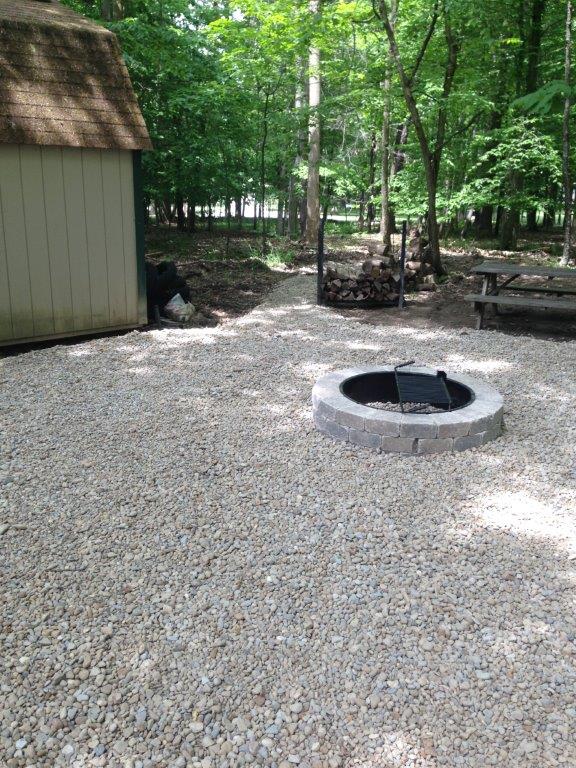 Gravel campsite with a circular fire pit beside a shed, surrounded by trees and picnic tables.